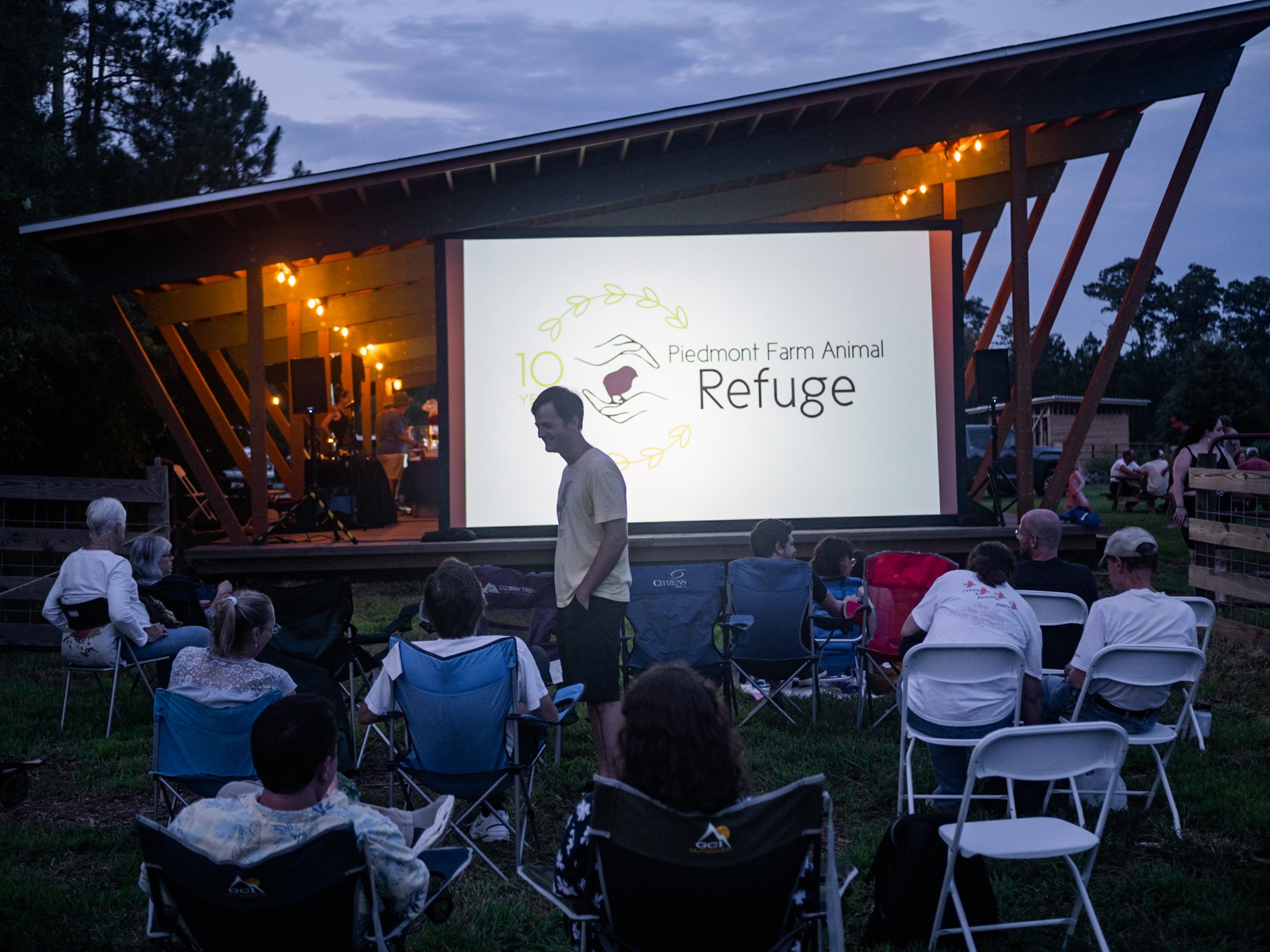 Audience watching an outdoor movie screening on a large screen at dusk, with trees and sky in the background, event celebrating Piedmont Farm Animal Refuge's 10th anniversary.