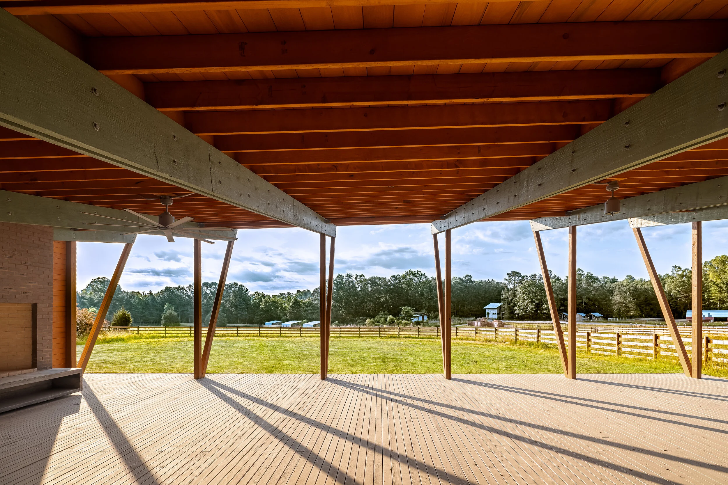 View of a covered outdoor patio with wooden beams and ceiling overlooking a green field and trees in the distance.