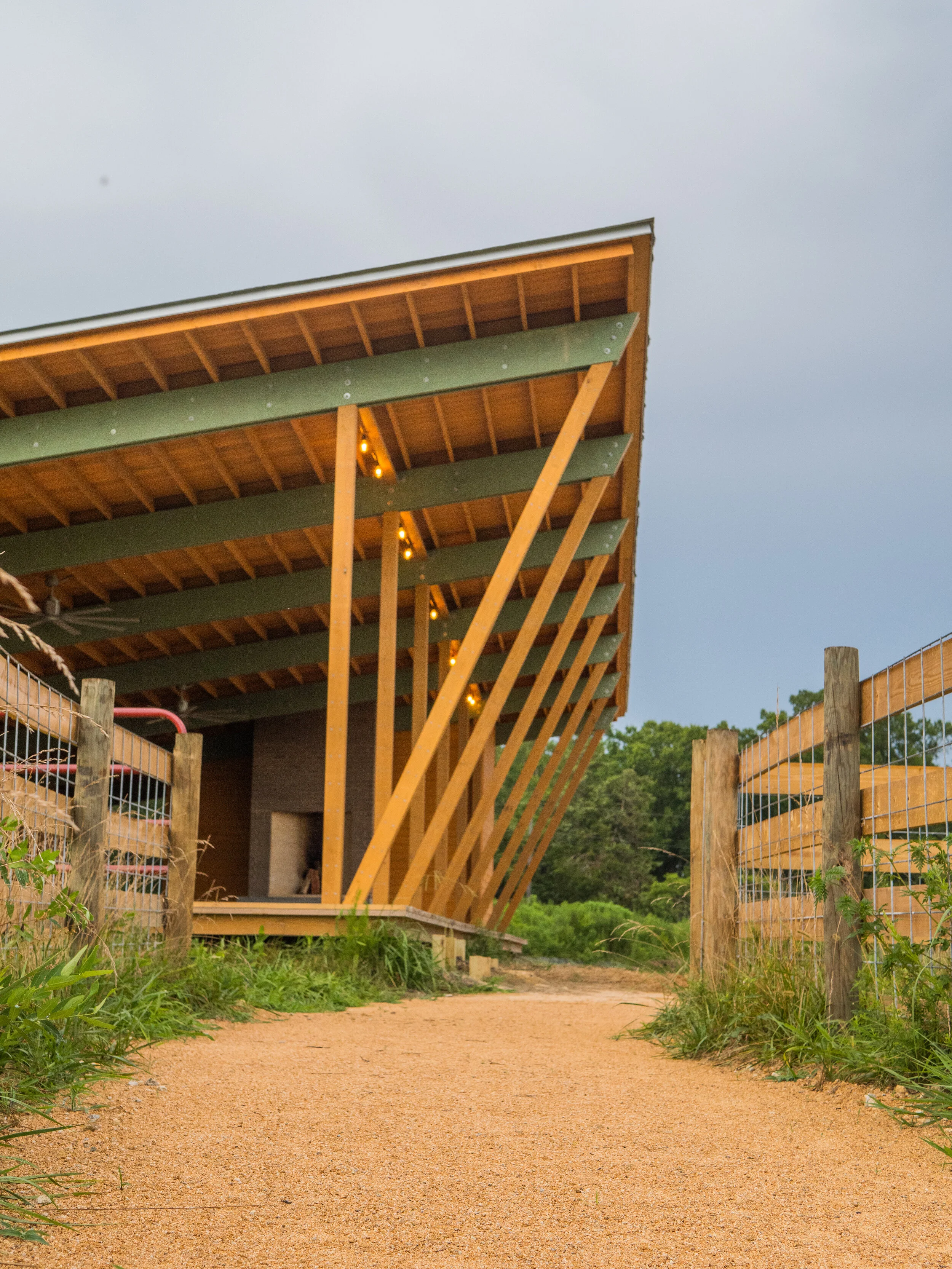 A building under construction with exposed wooden beams, a slanted roof, and string lights. A dirt pathway leads towards the building, surrounded by green grass and fencing on both sides.