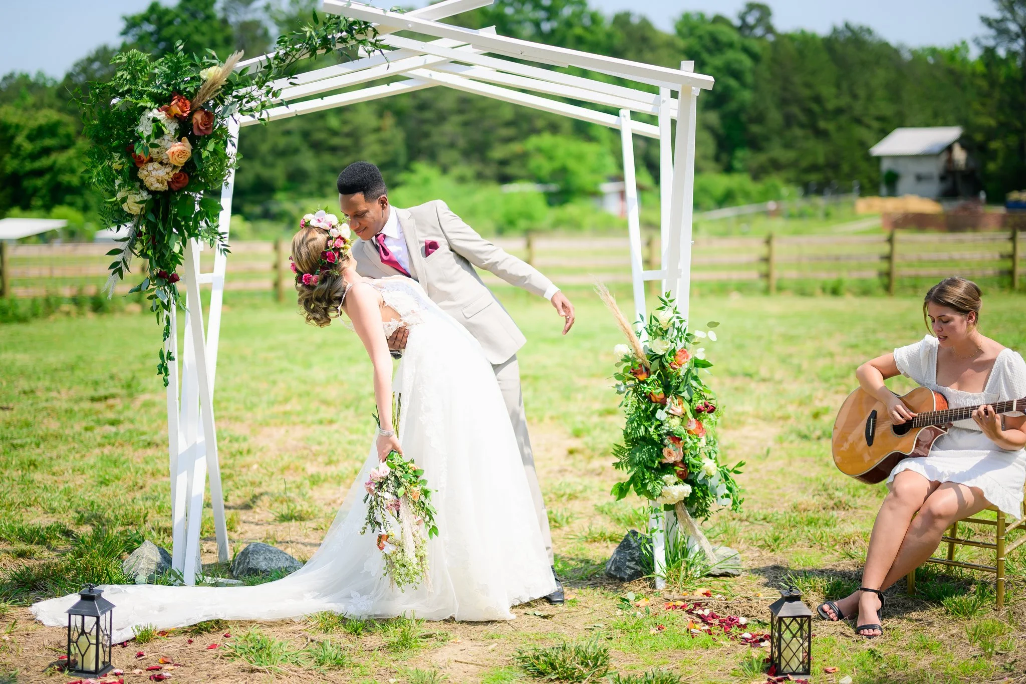 A bride and groom sharing a romantic moment under a decorated wedding arch outdoors, with a musician playing guitar beside them.