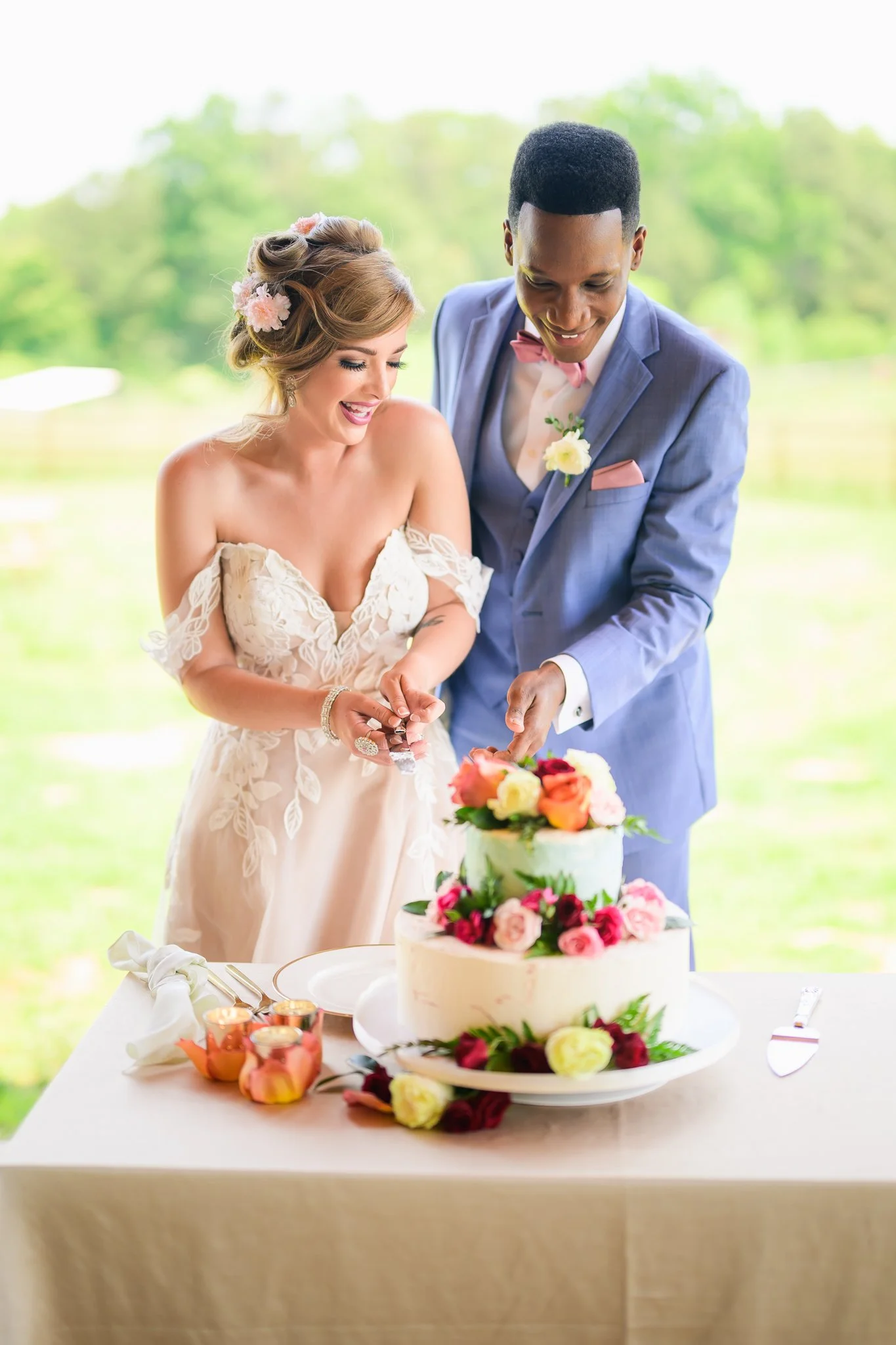 A bride and groom cutting a wedding cake decorated with pink, red, yellow, and white flowers during their outdoor wedding reception.