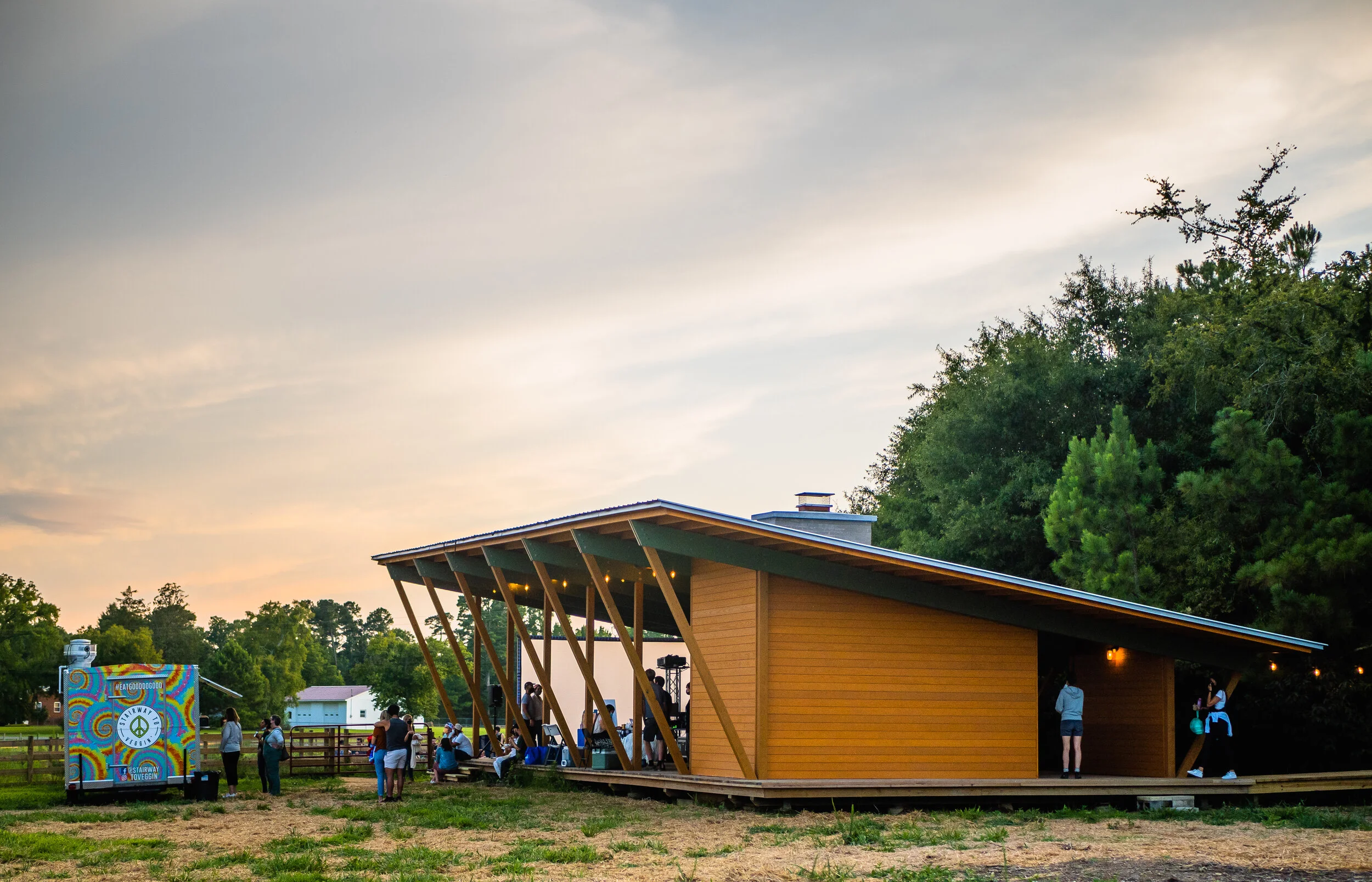 People gathered outside a modern wooden building with an open-air stage during sunset. A colorful sign in the foreground features peace symbols and the words 'Stairway to Vegan.'