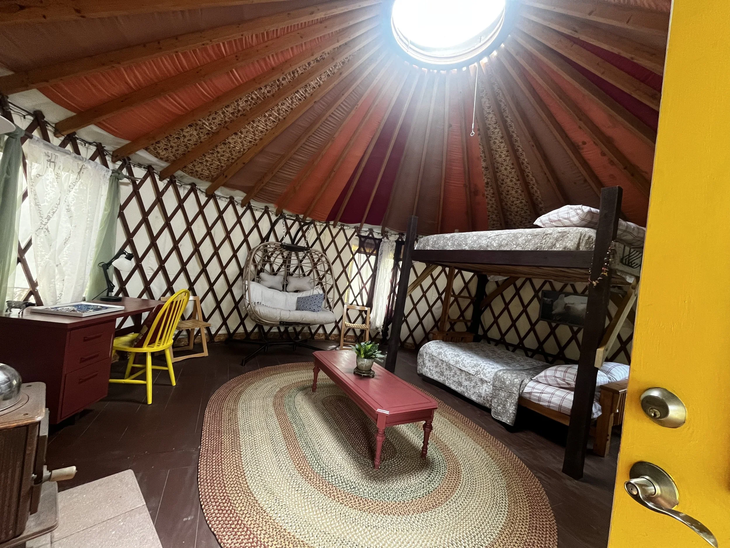 Interior of a yurt showing wooden walls, a circular skylight, various furniture including a bed, a wicker chair, a red table, a yellow chair, and curtains on the windows. The floor has a braided rug.