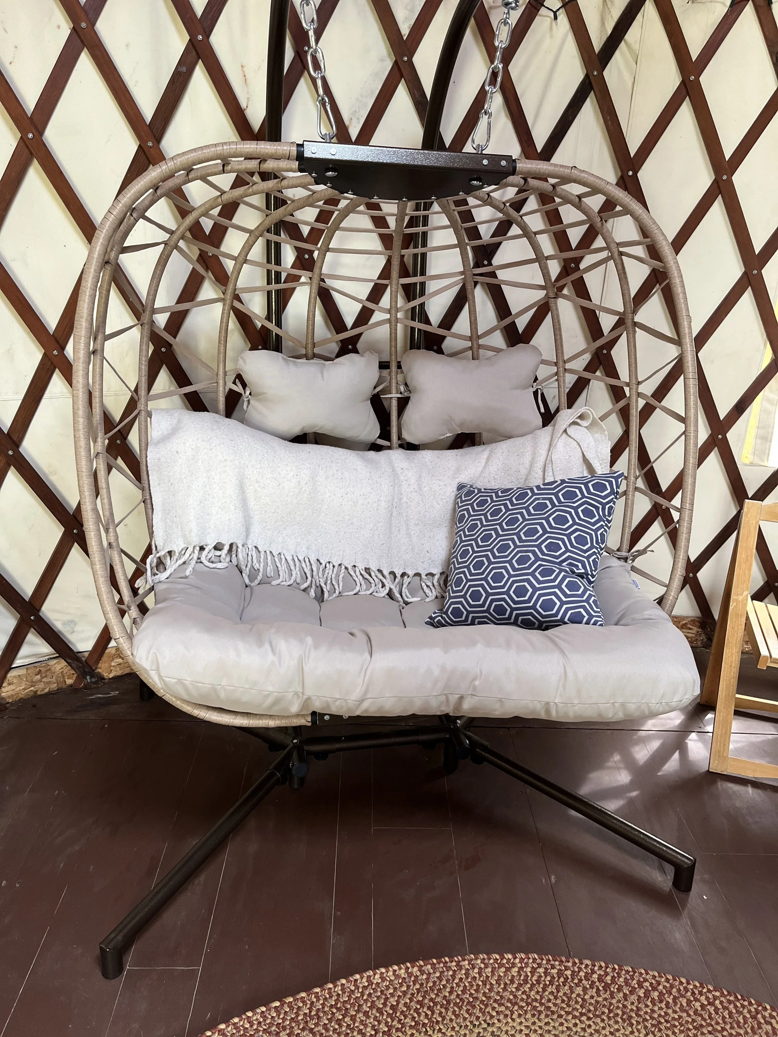 A hanging chair with a cushioned seat and two pillows, set against a wooden lattice wall. The chair has a black metal frame and is suspended by chains.