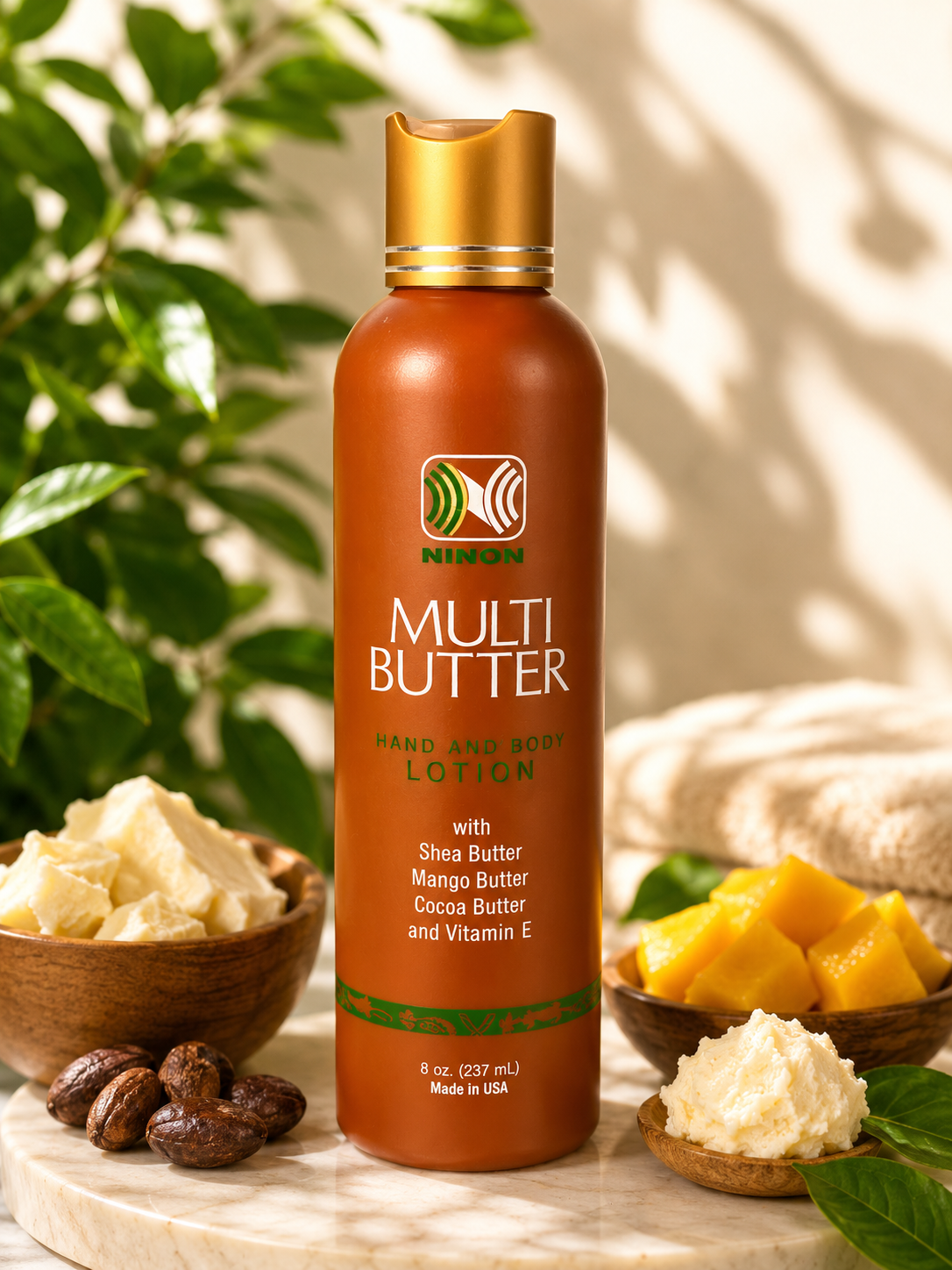 An orange bottle of Ninon Multi Butter Hand and Body Lotion with Shea Butter, Mango Butter, Cocoa Butter, and Vitamin E, surrounded by bowls of shea butter, mango chunks, and a beige towel, with green leaves in the background.