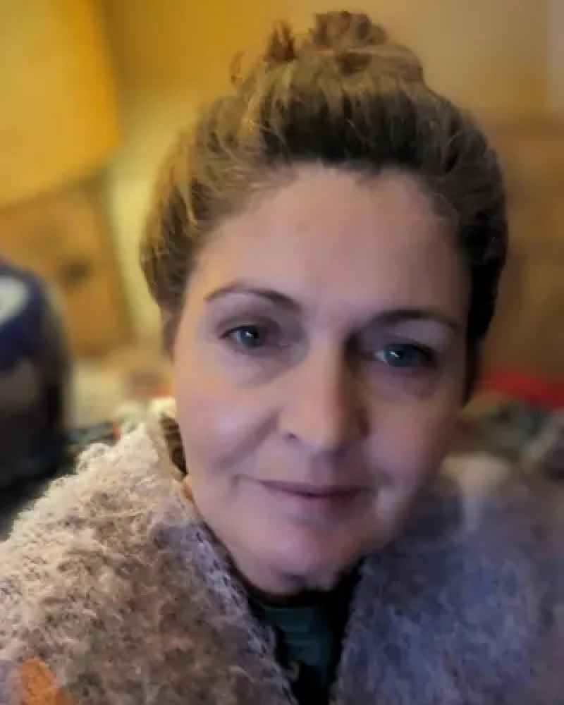 A woman with light skin, brown hair pulled back, and blue eyes, smiling at the camera indoors with a wooden background.