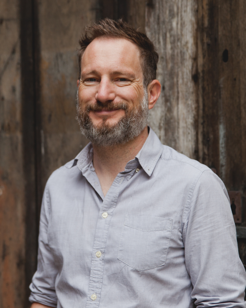 A man with short brown hair and a beard, smiling, wearing a light gray button-up shirt, standing in front of a rustic wooden wall.