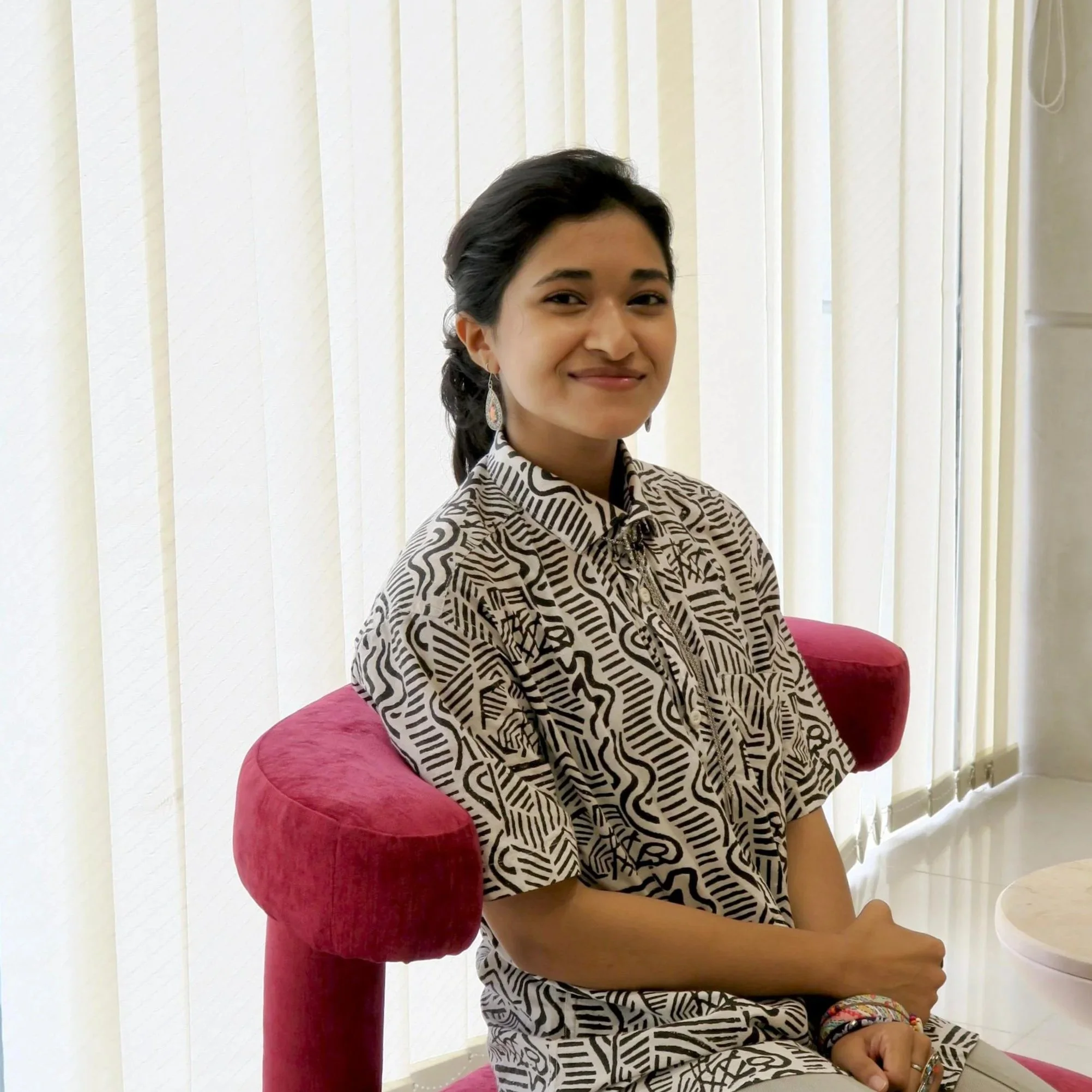 A young woman with dark hair styled in loose waves, wearing a black and white patterned dress, sitting on a red cushioned chair with white vertical blinds behind her, smiling gently at the camera.