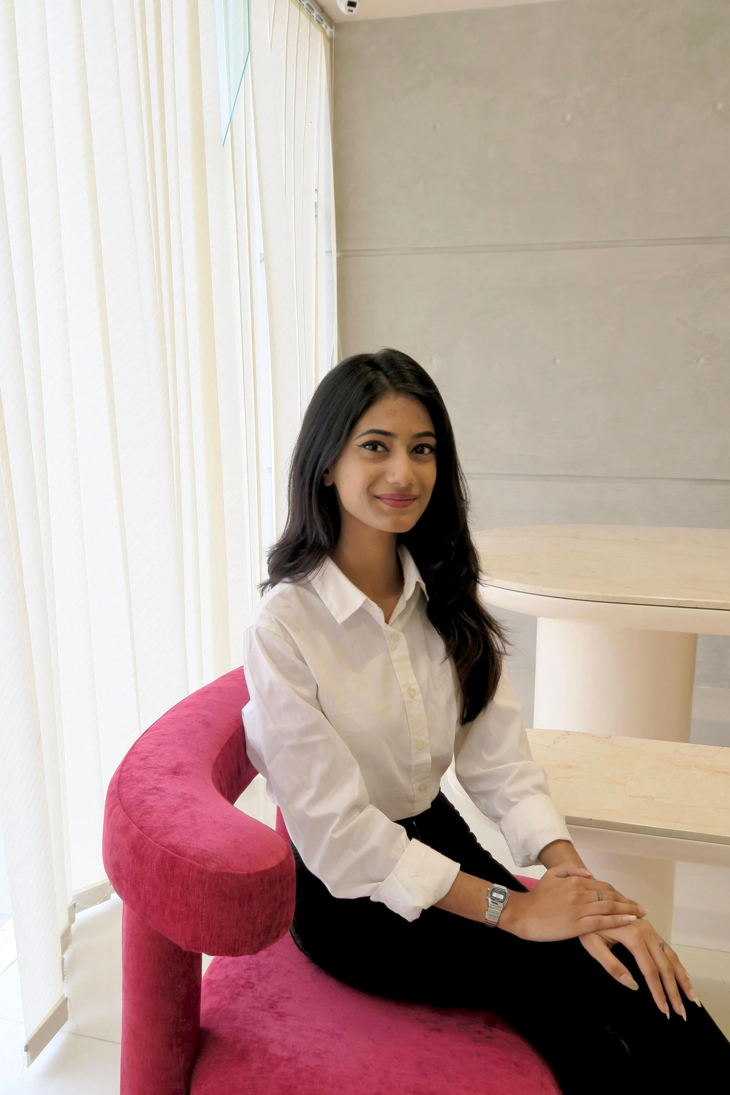A young woman with long dark hair sitting on a pink velvet chair near a window with vertical blinds, in a room with light-colored walls, wearing a white button-up shirt and a wristwatch.