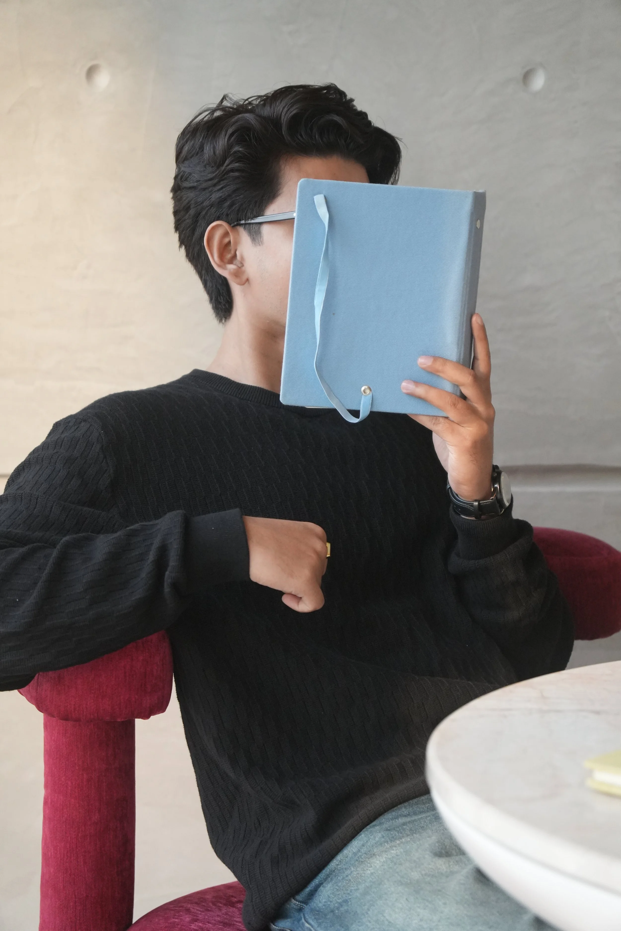 A person sitting on a red chair, holding a blue book or folder in front of their face, with their elbow resting on the armrest and their other hand near their waist. They are wearing a black sweater, a wristwatch, and glasses, with short dark hair. The background features a light-colored wall.