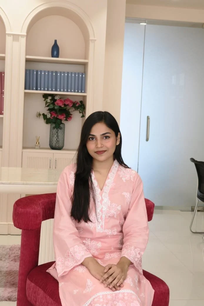 A woman in a pink embroidered kurta sitting on a red velvet chair indoors with a white bookshelf behind her, decorated with flowers and a blue vase.