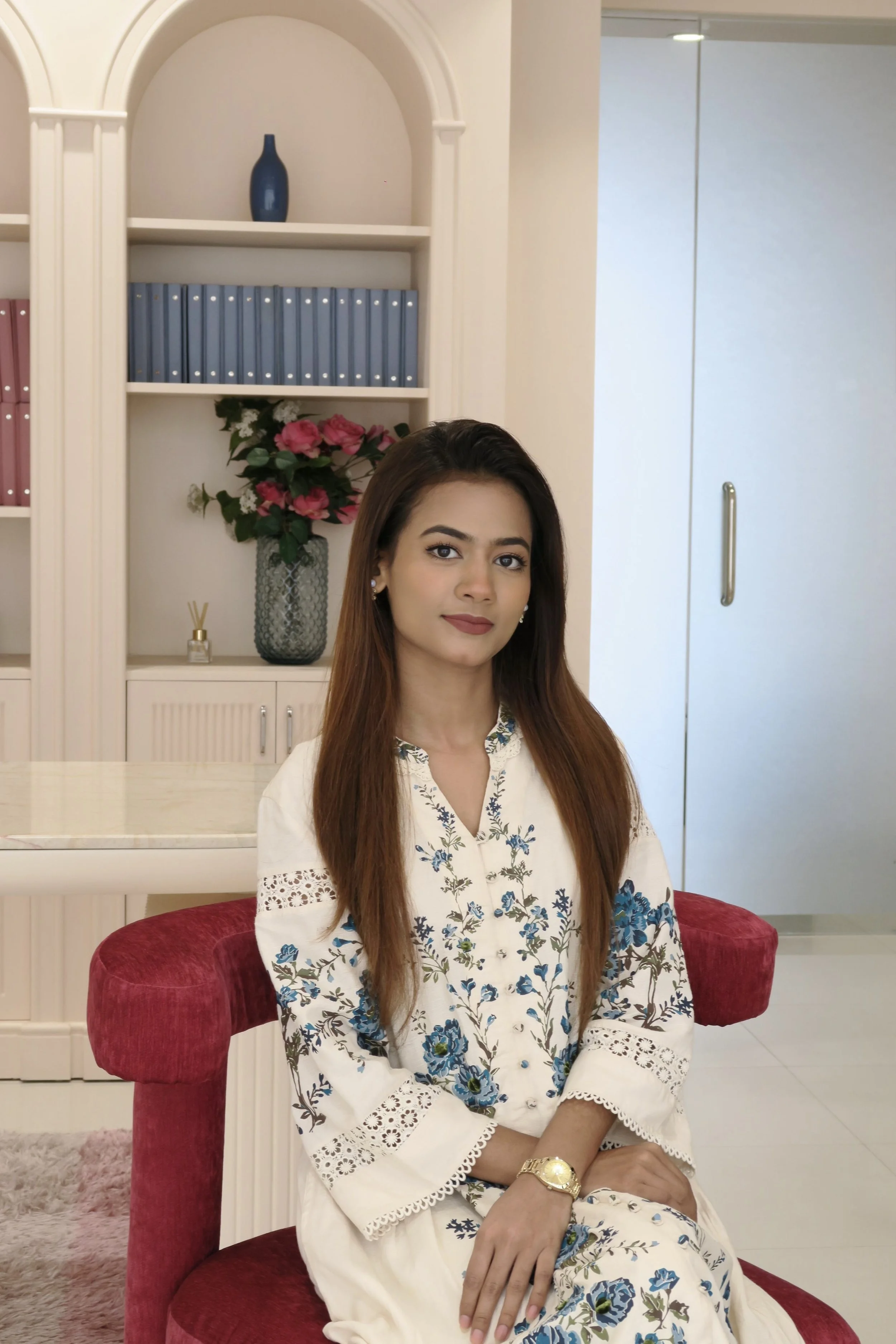 A young woman with long brown hair, wearing a white dress with blue floral embroidery, sitting on a red velvet chair in a room with elegant decor. She has a gold watch on her left wrist and pearl earrings.