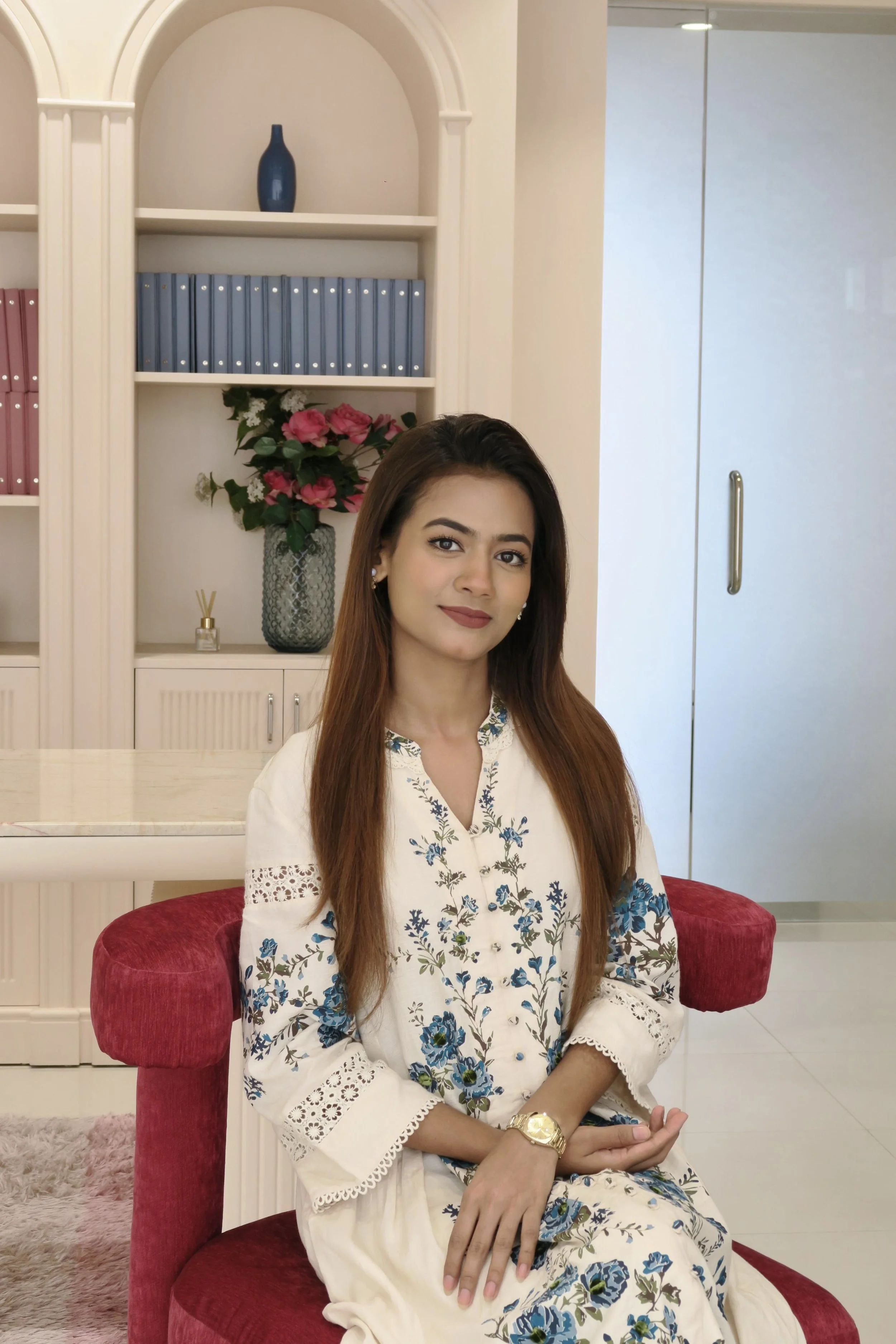A young woman with long brown hair, wearing a white dress with blue floral embroidery, sitting on a red velvet chair in a room with elegant decor. She has a gold watch on her left wrist and pearl earrings.