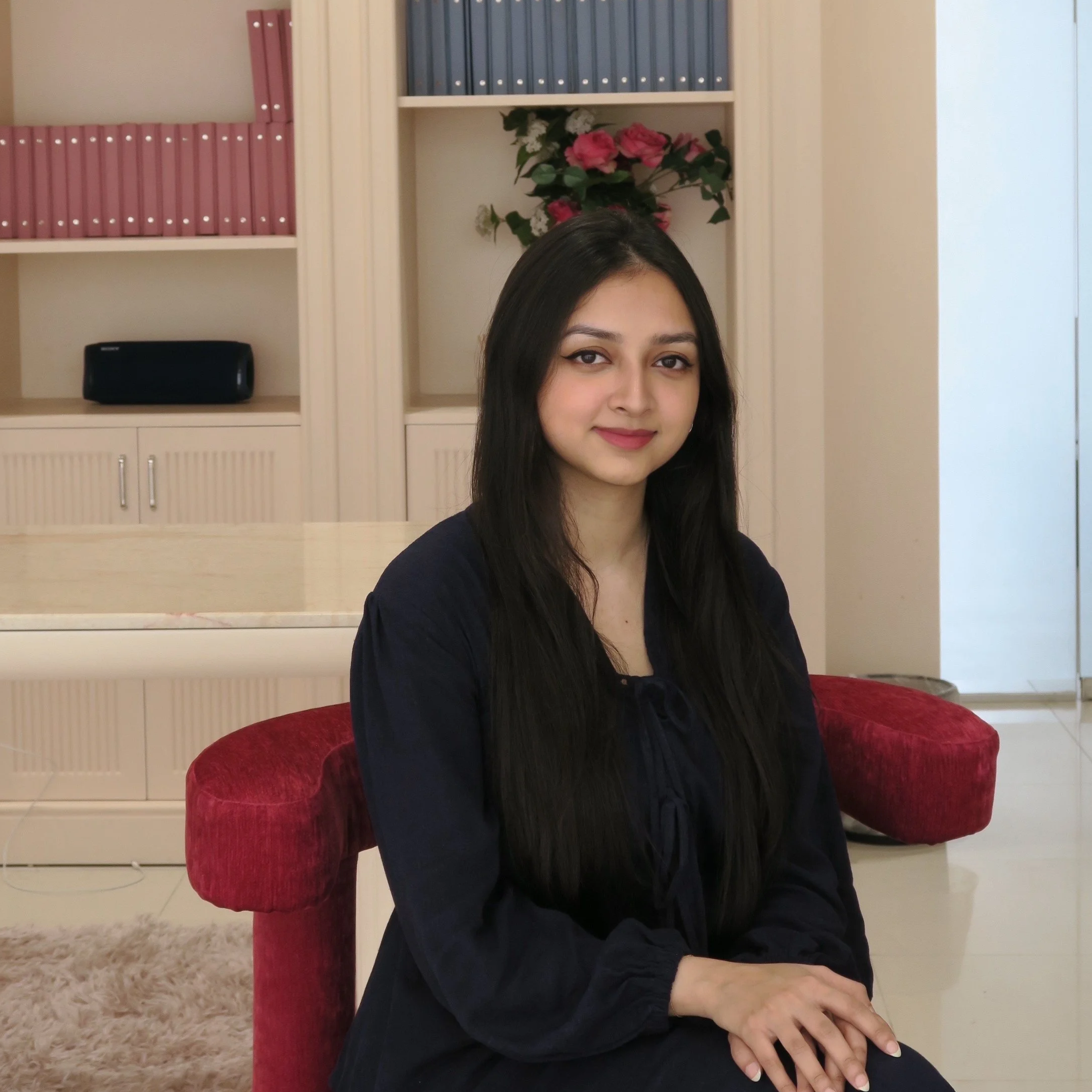 A young woman with long black hair, wearing a dark top, sitting on a red armchair in a room with beige walls, a white bookshelf with pink and blue books, a speaker, and a floral arrangement in the background.