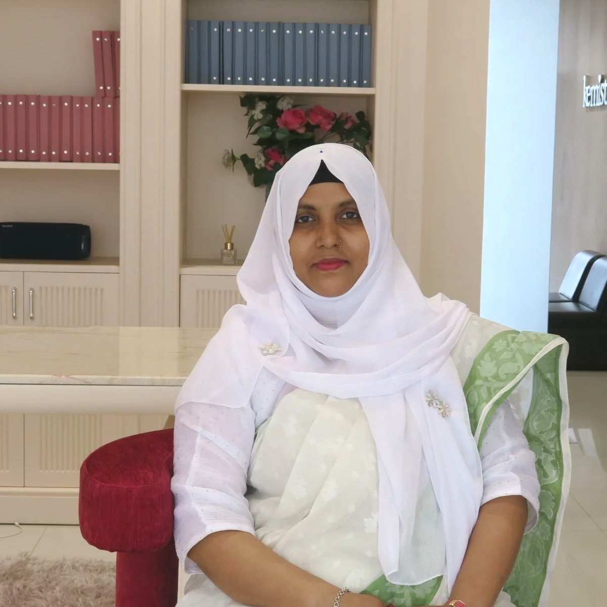 A woman in white traditional attire with a white head covering, sitting indoors in a modern office or reception area with bookshelves, pink flowers, and a black speaker in the background.