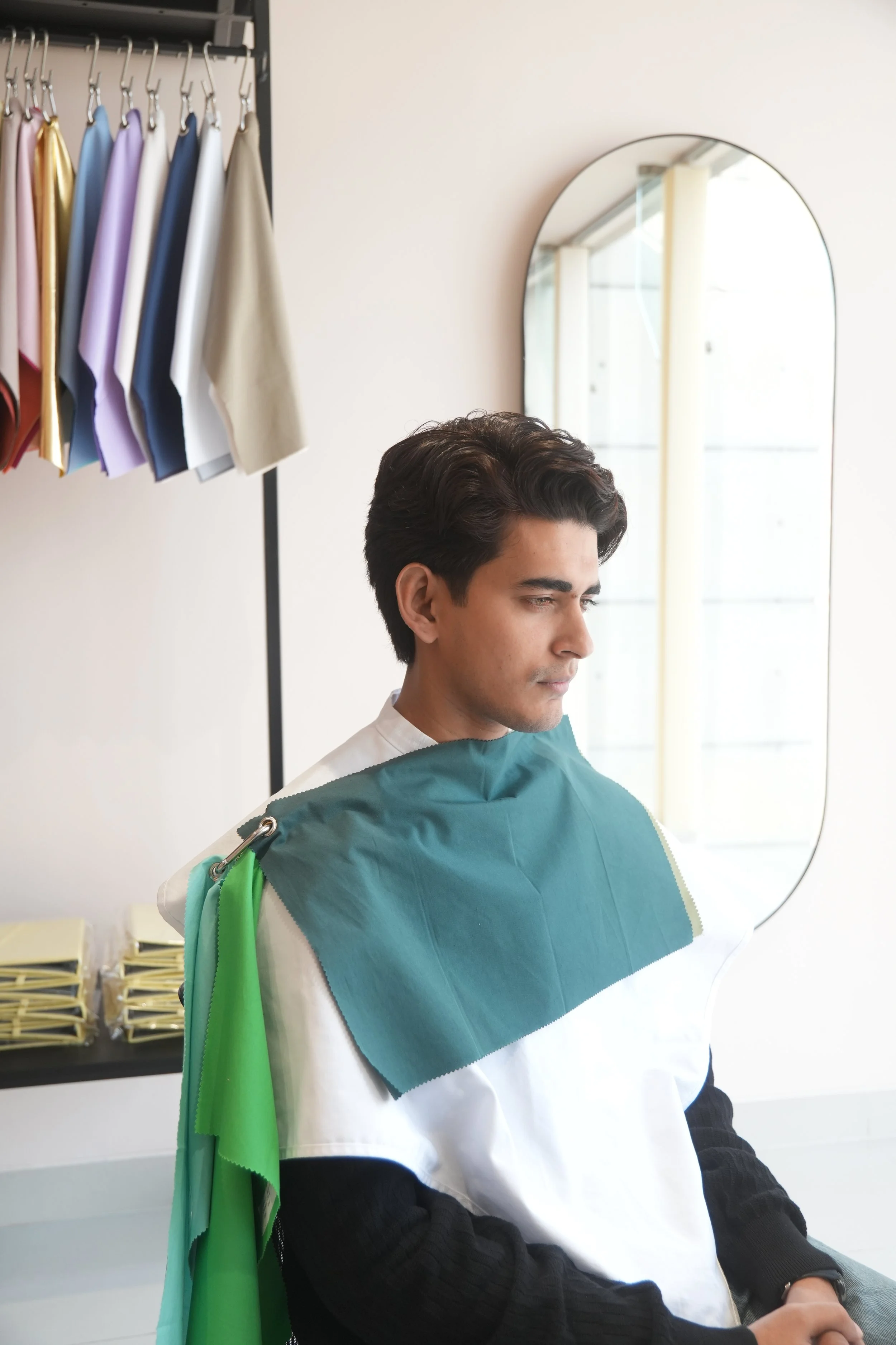A man sitting in a salon chair, preparing for a haircut, with a green cape draped over his shoulders, in front of a mirror with colorful cloth swatches hanging in the background.