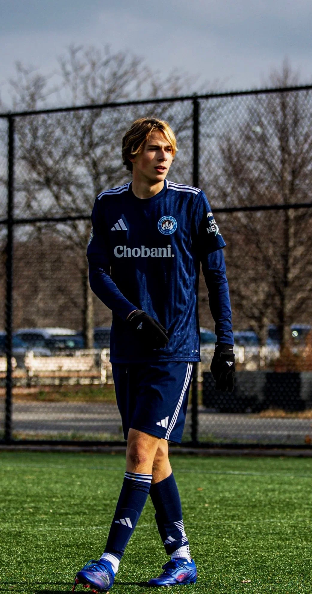Johnny Sullivan MLS Next Soccer player on homegrown academy, A young man in a navy blue soccer uniform standing on a soccer field, with a black chain-link fence and leafless trees in the background.