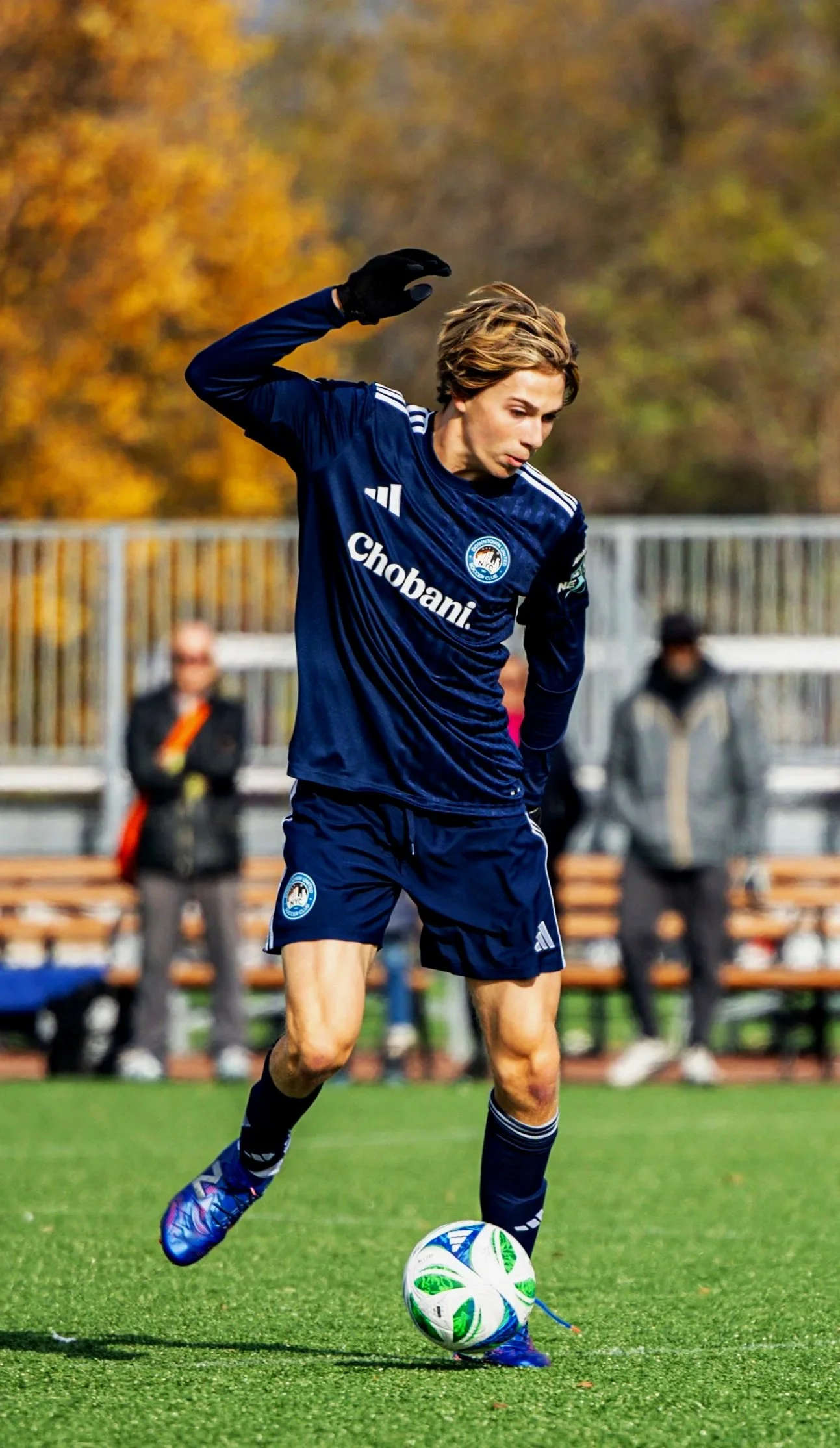 Johnny Sullivan the athlete showing soccer skills, A young male soccer player in blue uniform kicking a soccer ball during a game on a green field with autumn trees in the background.