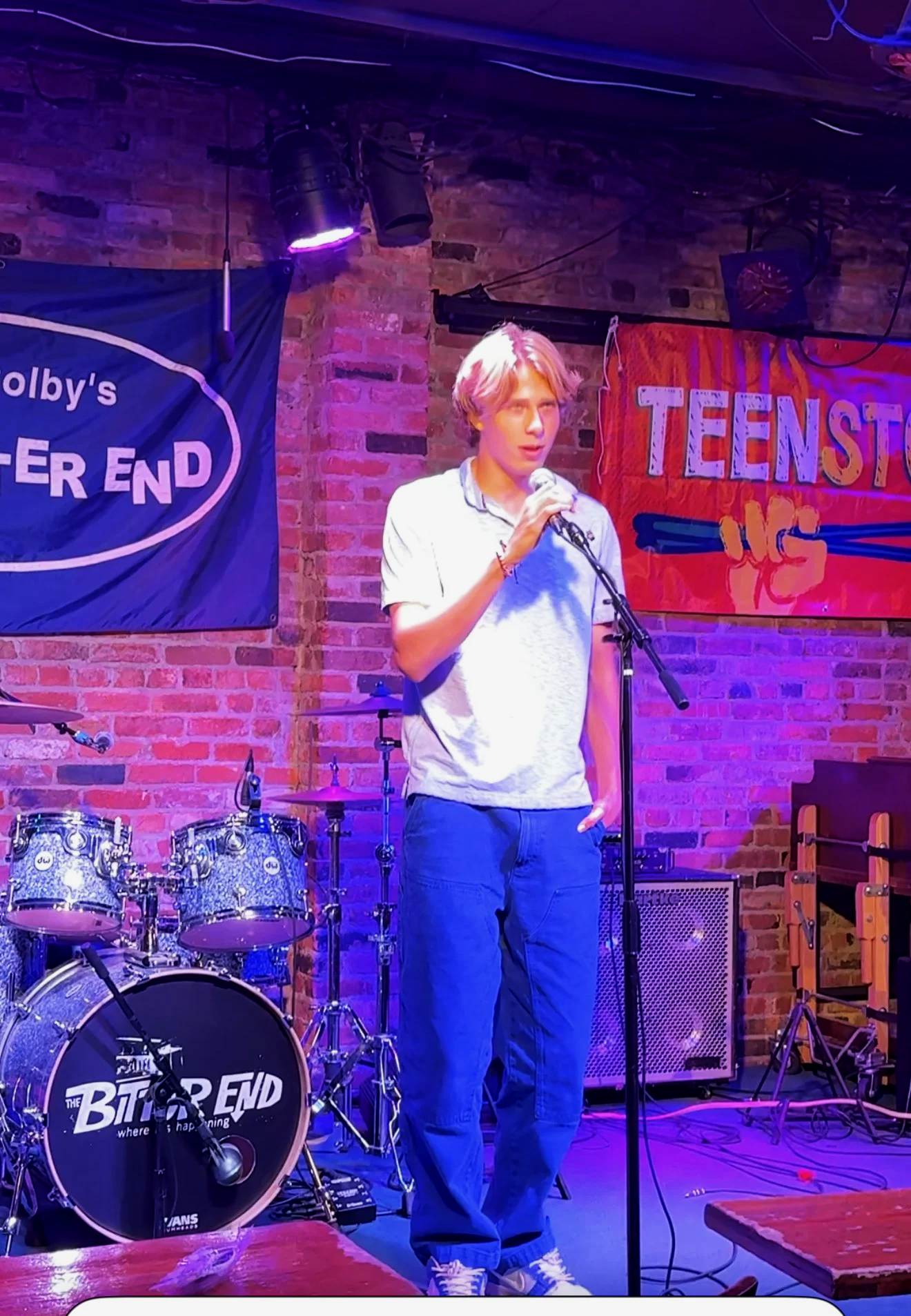 A young man with blond hair holding a microphone, standing on a stage with a brick wall background, music instruments, and banners that say 'Teenstage N' and 'The Bitter End'.