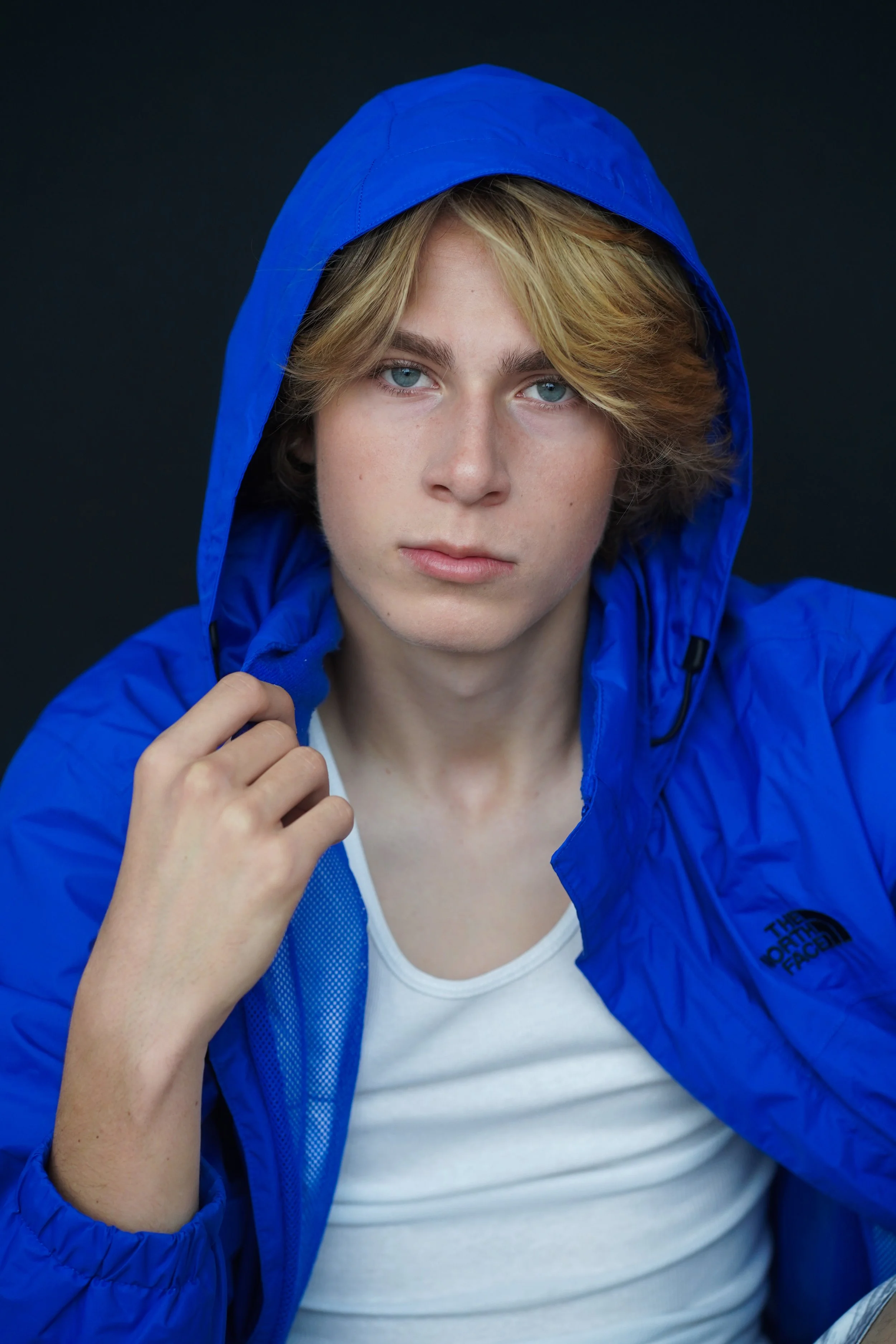 Johnny Sullivan, pop star, singer, with blond hair and blue eyes wearing a blue hooded jacket and a white shirt, looking directly at the camera against a dark background.