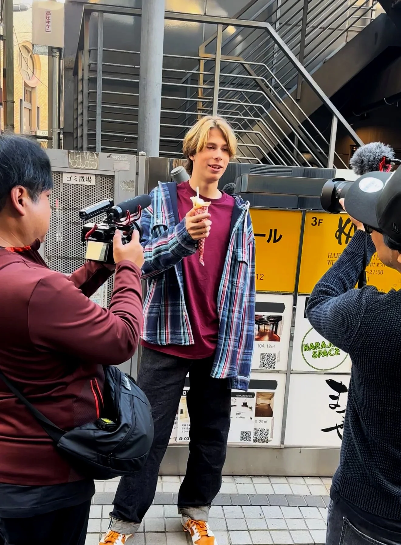 Johnny in Japan being filmed in Tokyo after winning the national singing contest and being offered a record label deal. A young man holding an ice cream cone being filmed by two cameramen in an urban setting with stairs and signage in the background.