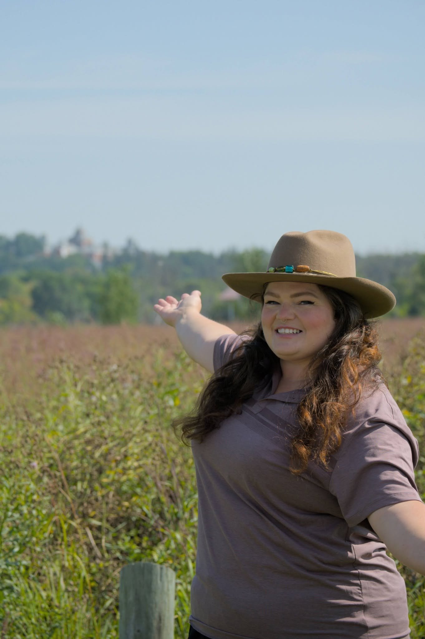 Tour Guide and owner, Alison, A curly-haired woman in a wide brim hat, is showing the scenery in Southwestern Manitoba. An expanse of greenery is behind her.