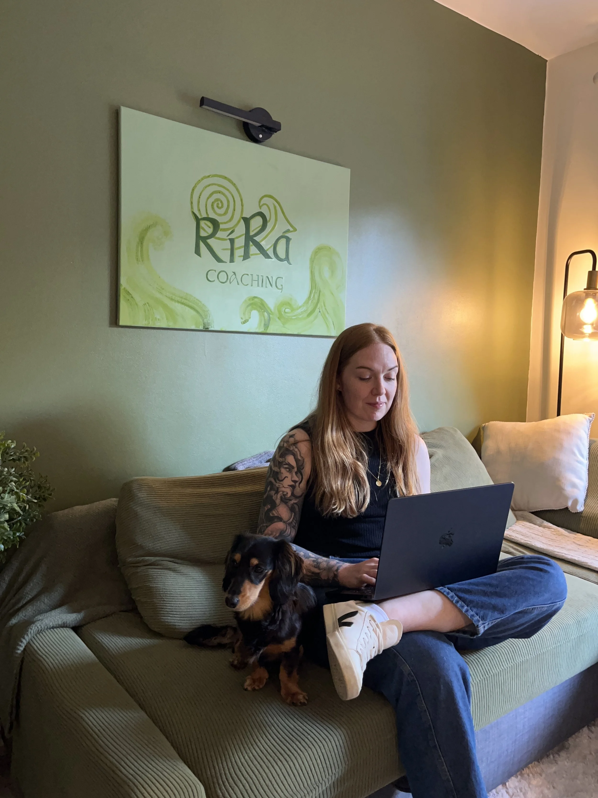 A woman with long red hair and tattoos on her arm is sitting cross-legged on a green couch, working on a laptop while a small black and brown dog sits beside her. The room has green walls, a lamp on a stand, and a wall art sign that says 'Rira Coaching' with a green swirl design.