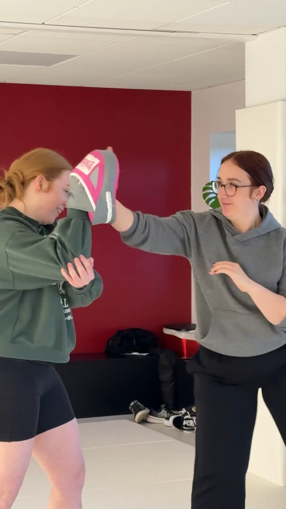 Two women are practicing self-defense techniques in a room with a red wall. One woman is blocking a punch from the other woman, who has a gray and pink glove on her hand. Sneakers are visible on the floor in the background.