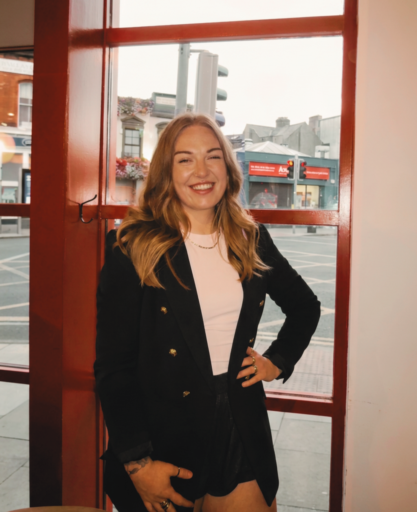 The coach, Ríana. A smiling woman with wavy, shoulder-length hair standing inside a building near a window and red door frame, with an urban street scene visible outside.