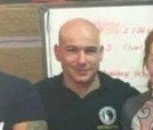 A man with a shaved head smiling, wearing a dark shirt with a logo, standing indoors near a whiteboard.
