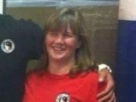 A smiling woman with brown hair and bangs, wearing a red sports jersey, standing next to someone in a dark shirt, in a sports setting.