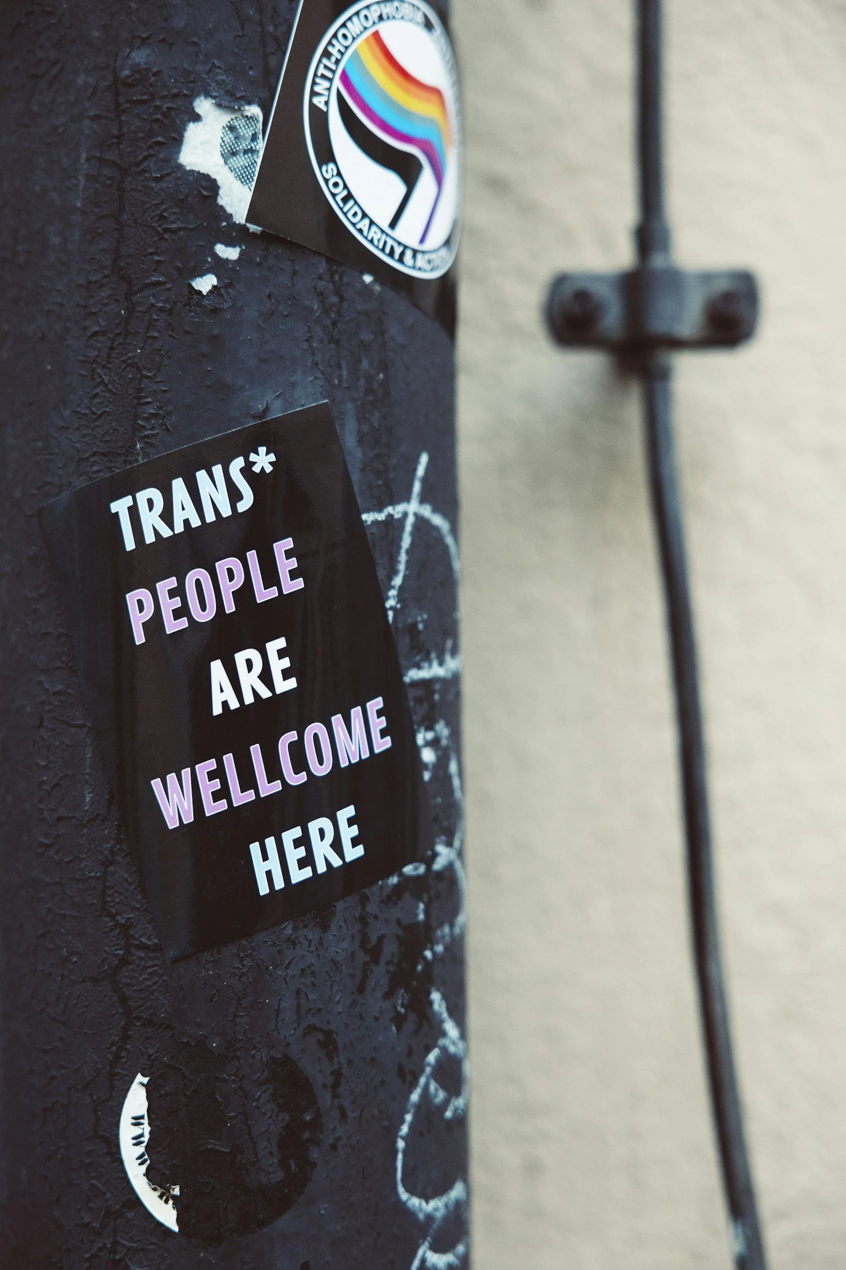 Close-up of a black pole or post with a sticker that reads 'Trans* People Are Welcome Here' in white and pink text. The pole has some worn paint and additional stickers, and there's a beige wall and black electrical conduit in the background.