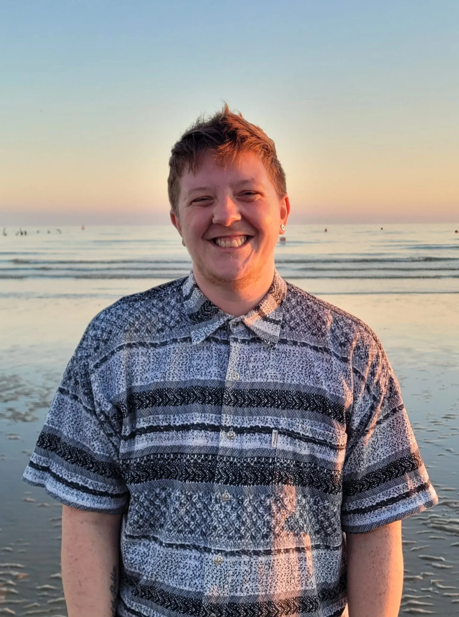 A smiling young man with short hair and earrings standing on a beach during sunset, wearing a patterned button-up shirt. The ocean and a few buoys are visible in the background.