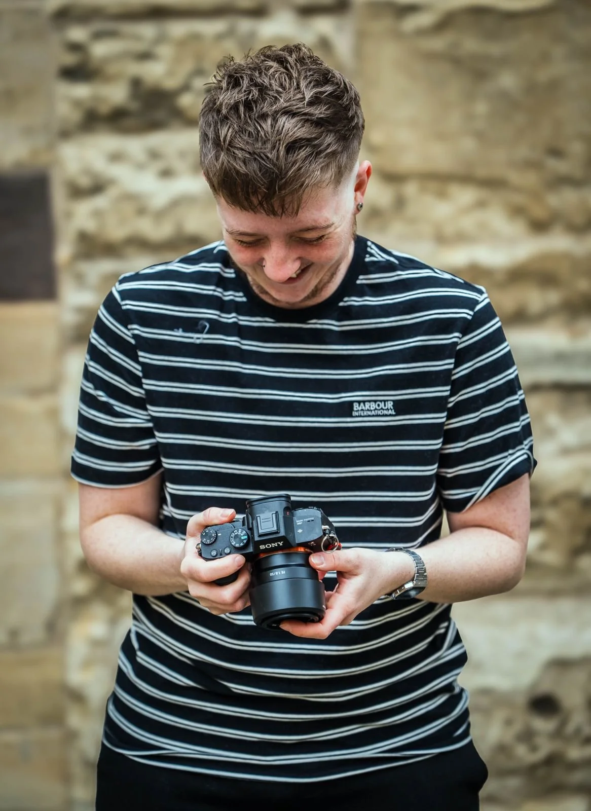 A young man with curly hair, smiling and looking down at a Sony camera he is holding, standing in front of a beige stone wall.