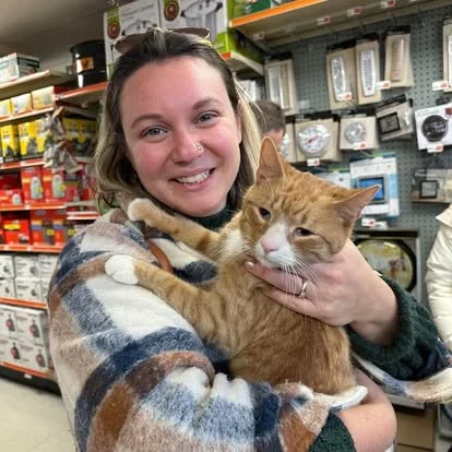 A woman smiling and holding an orange tabby cat in a store aisle with pet supplies.
