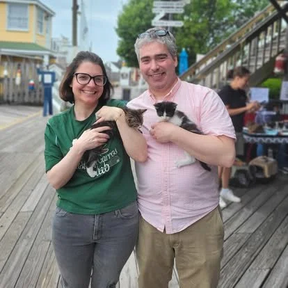 A man and woman standing outdoors on a wooden deck, each holding a small kitten. The woman is wearing glasses and a green shirt, and the man is in a pink shirt. There are people and buildings in the background.