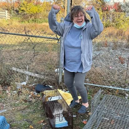 A woman standing outdoors near a chain-link fence, smiling and flexing her arms, with a cat in a cage and outdoor items around her.