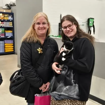 Two women, one older and one younger, standing indoors, holding a black and white puppy, smiling at the camera.