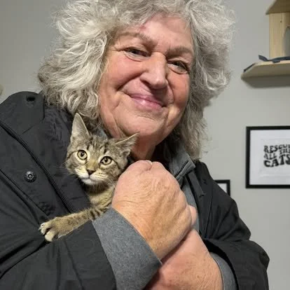 A smiling elderly woman with gray curly hair holding a small tabby kitten close to her chest in an indoor setting.