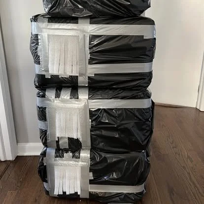 Stack of black plastic bags sealed with white packing tape, placed on a wooden floor near a white wall.