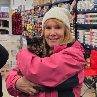 A woman in a pink jacket and white beanie holding a cat inside a store aisle.