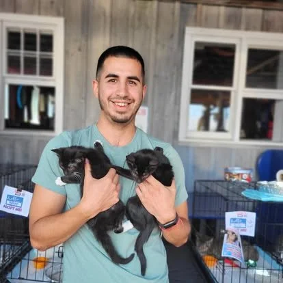 A young man with a beard smiling and holding two black puppies in front of animal cages in an indoor setting.