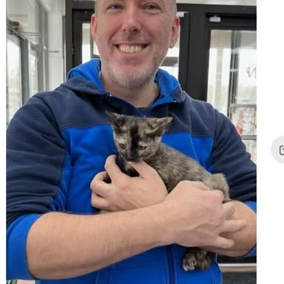 A man smiling and holding a small gray and black kitten indoors near glass doors.