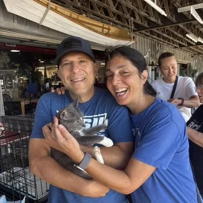 Two smiling people holding a gray and white kitten at an indoor event with a woman in the background.