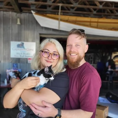 A smiling woman with blond hair and glasses holding a black and white kitten, standing next to a smiling man with a beard, inside a rustic building with a kayak hanging from the ceiling.