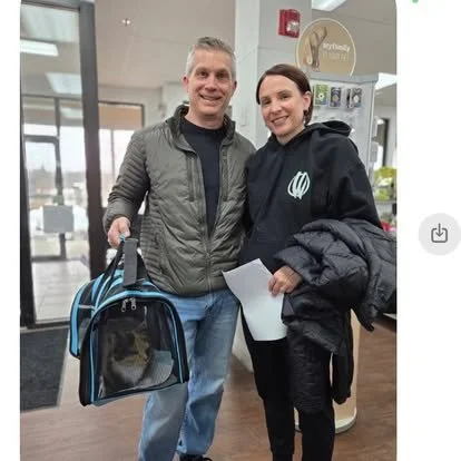 Two people, a man and a woman, standing together inside a building, smiling at the camera, with the man holding a pet carrier and the woman holding a piece of paper and a jacket.