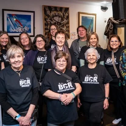 Group of ten women smiling, some wearing black BCR shirts, gathered in a room with framed pictures on the wall.