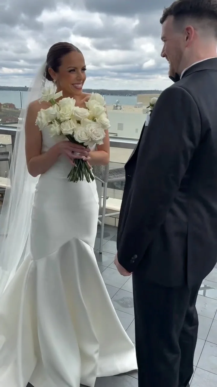 A bride holding a bouquet of white roses and smiling at a groom dressed in a black tuxedo during their wedding ceremony on a rooftop with a cityscape and water in the background.