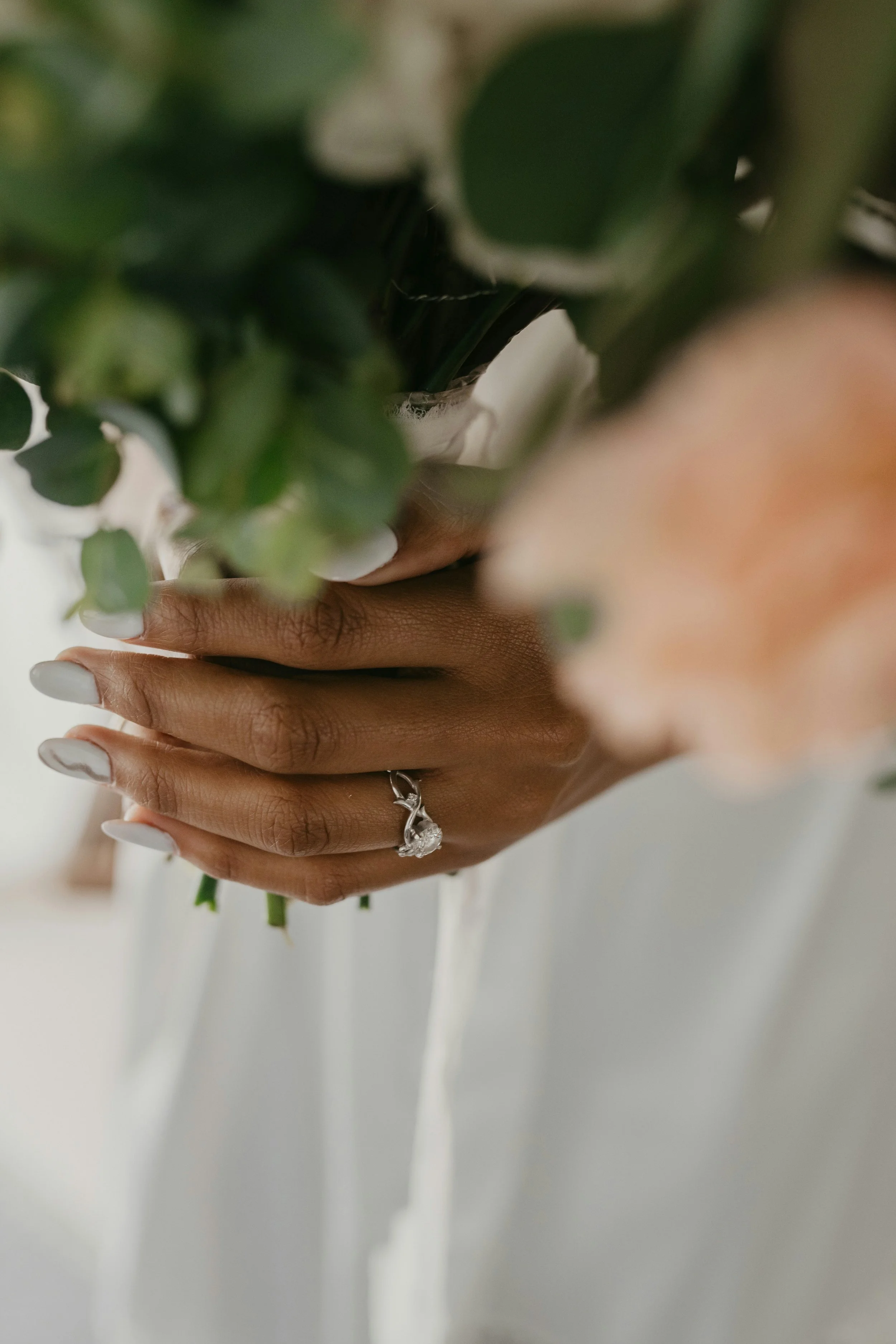 Close-up of a person's hand with a silver ring, holding a bouquet of green leaves and white flowers against a white background.