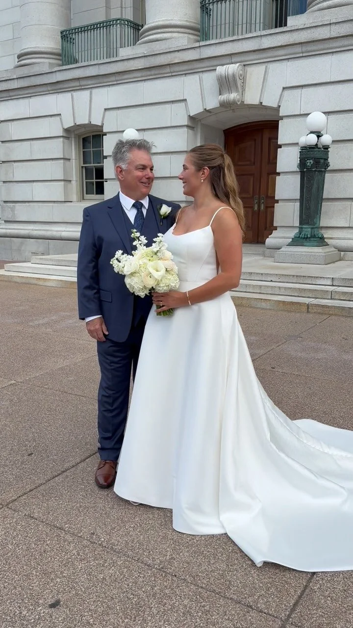 A bride and an older man, possibly her father, stand outside a historic building with white stone walls and steps. The bride wears a white wedding gown with a long train and holds a bouquet of white flowers. The man wears a navy blue suit with a white shirt and a tie. They are smiling and looking at each other.